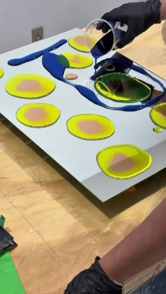 Mão com luva preta vertendo pigmento azul sobre um espelho com círculos amarelos.